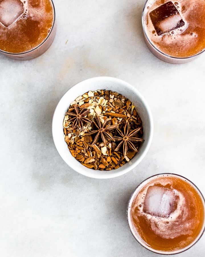 Three glasses of icy homemade root beer surrounds a small bowl filled with herbs like star anise, sarsaparilla, ginger, sassafras and licorice.