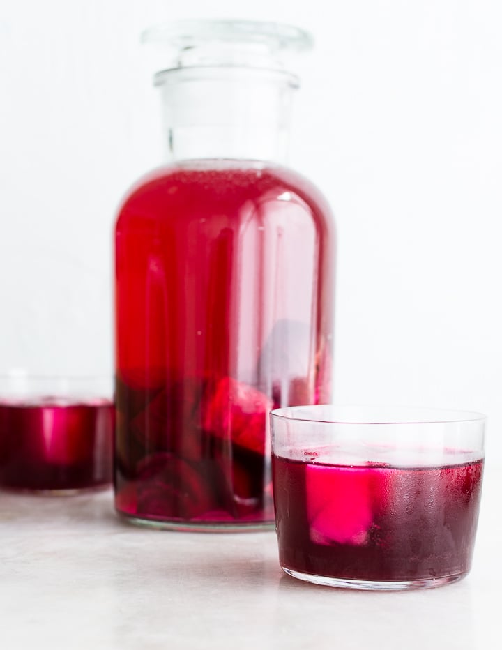 Two glasses of beet kvass next to an apothecary jar filled with beets fermenting in brine.