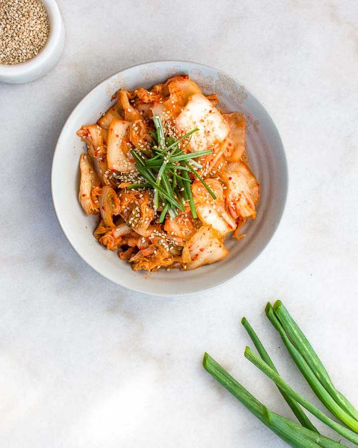 Homemade kimchi garnished by green onions and sesame seeds sits in an earthenware bowl.