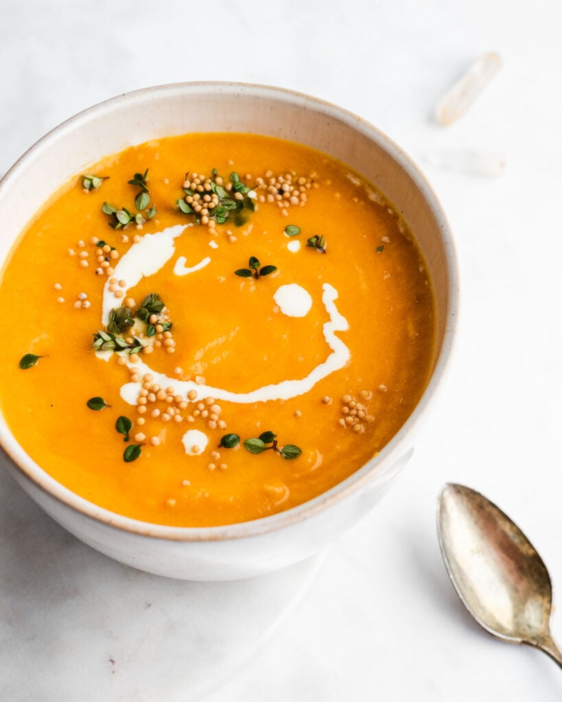 Carrot Leek soup, garnished with mustard seeds, thyme and sour cream, in an earthenware bowl.