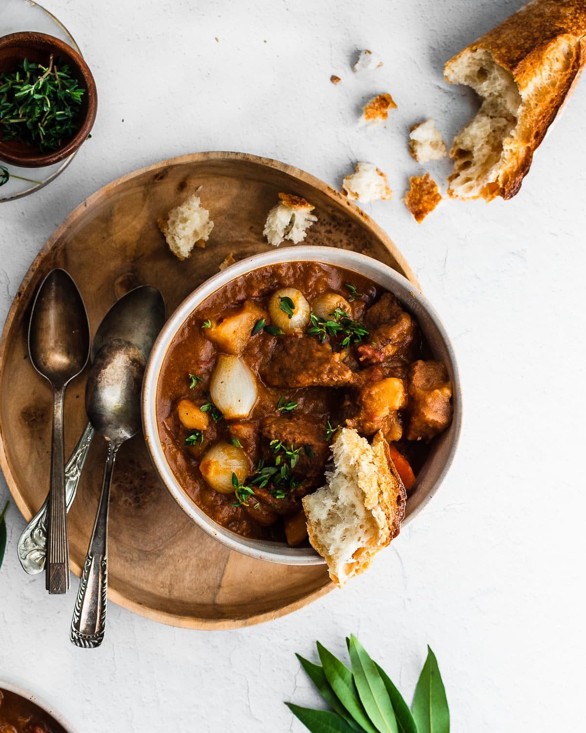 Beef and bacon stew in a bowl next to fresh thyme and a baguette