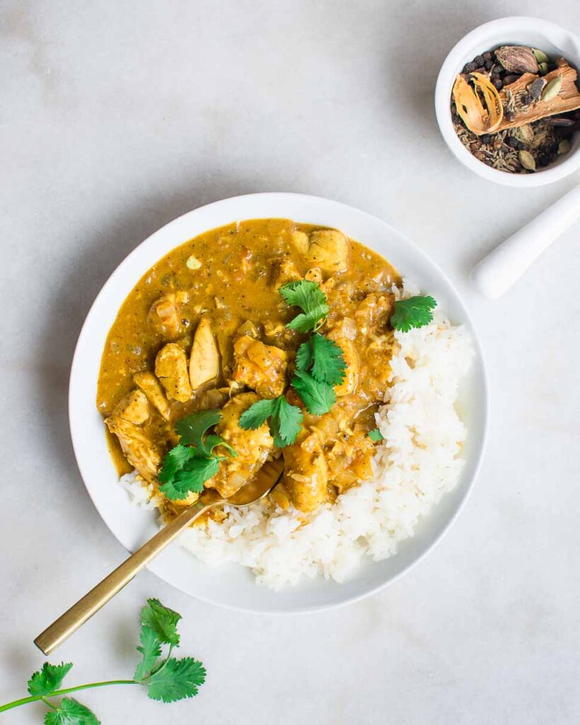 A serving of garam masala chicken curry sits in a white bowl, served over jasmine rice with cilantro. 