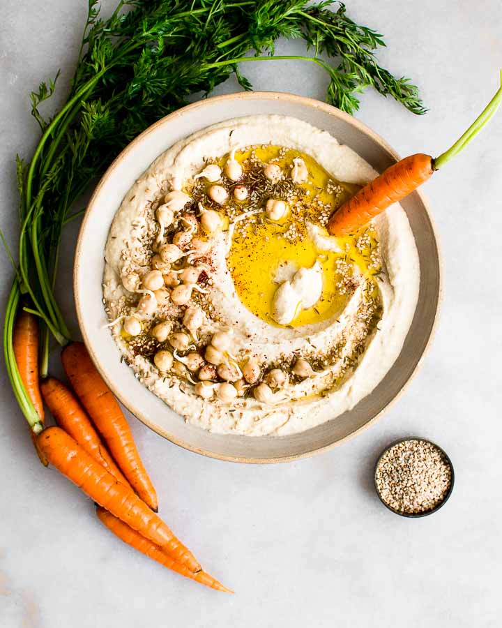 houmous germé dans un bol en terre cuite garni de pois chiches germés, de cumin et de graines de sésame et servi avec des carottes.