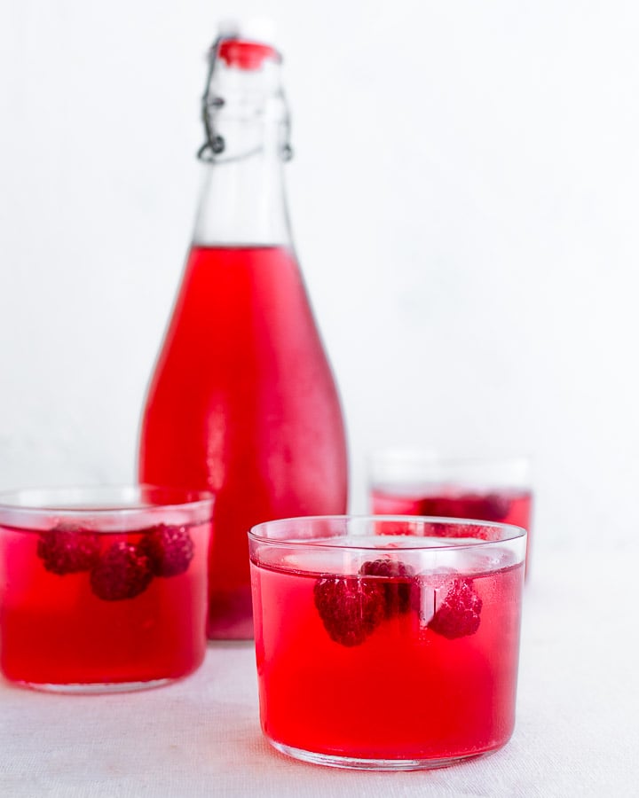 Fermented raspberry soda in a glass with raspberries and ice next to a bottle of raspberry soda.