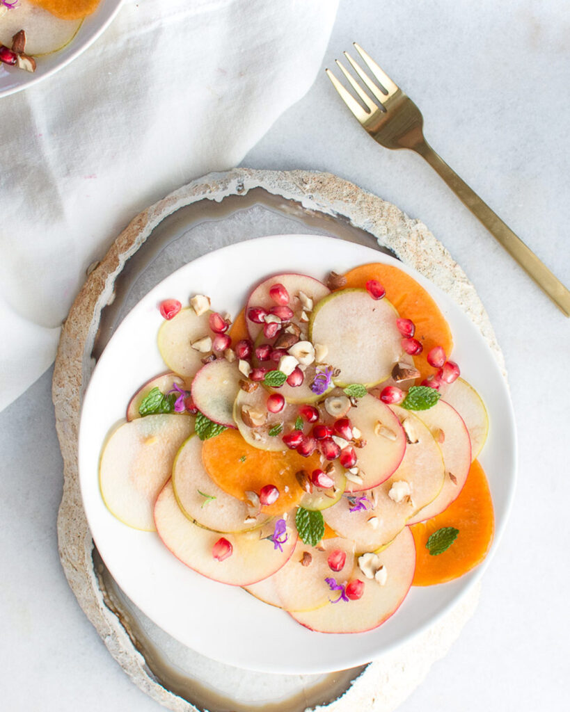 Autumn fruit salad with shaved apples, pears, and persimmons garnished with pomegranates and hazelnuts.