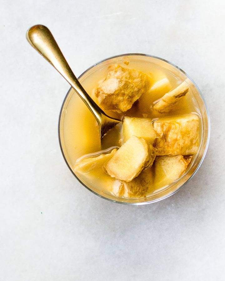 Chunks of fresh ginger sit in a glass of ginger bug on a marble tray with a gold spoon.