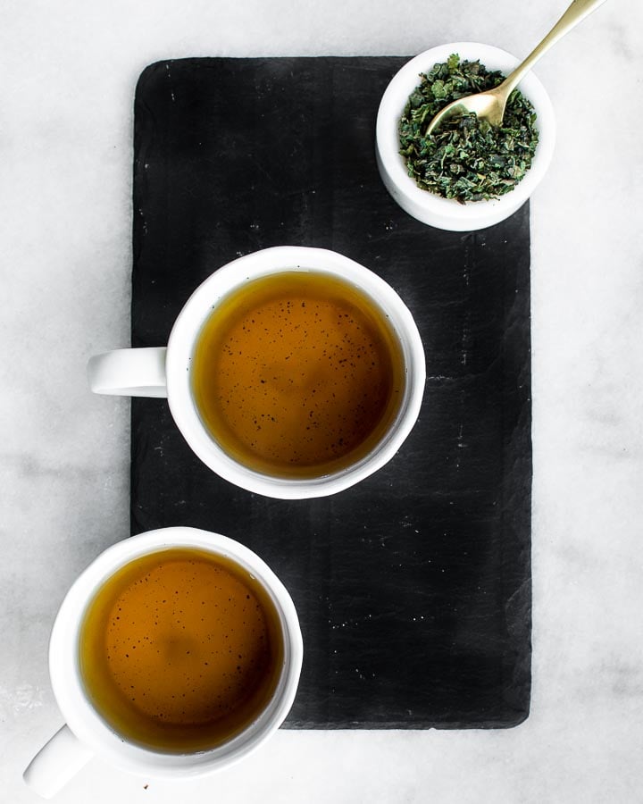 Two cups of nettle infusion on a slate board with a small dish full of dried stinging nettles.