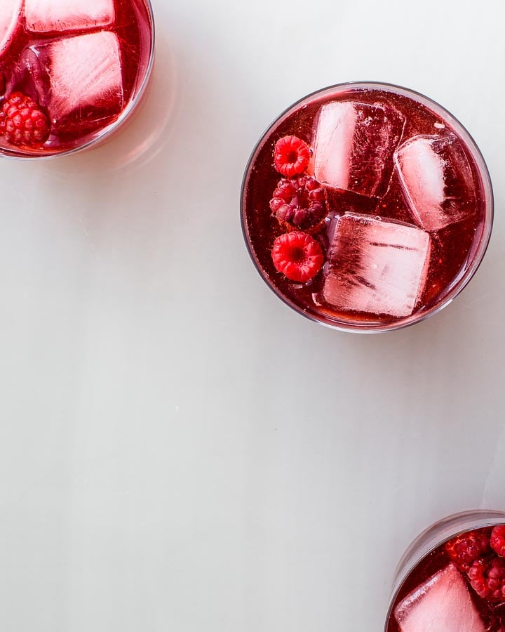 Three glasses of raspberry shrub served over ice, garnished with fresh raspberries.