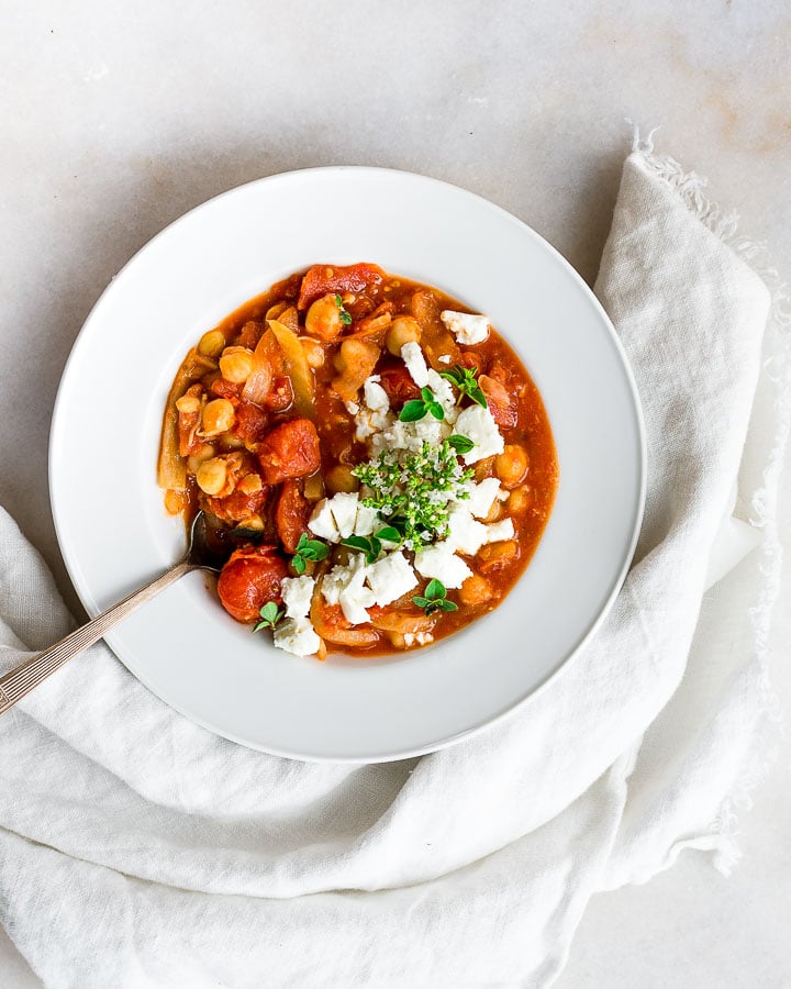 Chickpea stew with tomatoes, garnished with flowering oregano and crumbled feta cheese in a bowl on marble.