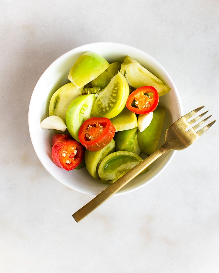 Fermented green tomatoes in a bowl with a fork on marble.