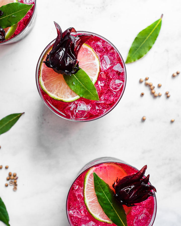 Hibiscus syrup poured into three glasses of ice, topped with lime, bay leaf and  preserved hibiscus flowers