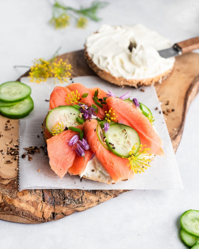 Sourdough bagel topped with cream cheese, cucumbers, chives, dill and lox on a wooden board.