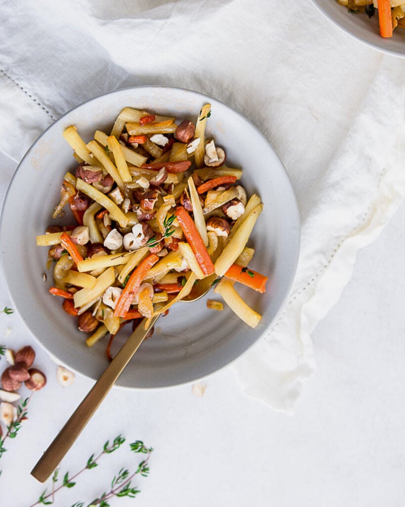 Maple-glazed root vegetables in a stoneware bowl garnished with thyme and hazelnuts.