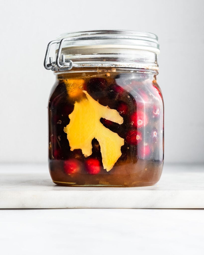 Fresh cranberries fermenting in a jar of honey with ginger, coriander, and orange peel