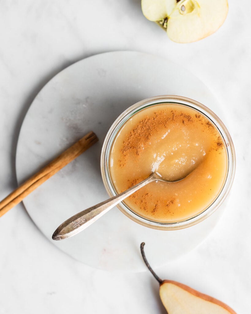 Pear applesauce in a glass bowl next to sliced pear, sliced apple, and a cinnamon stick.