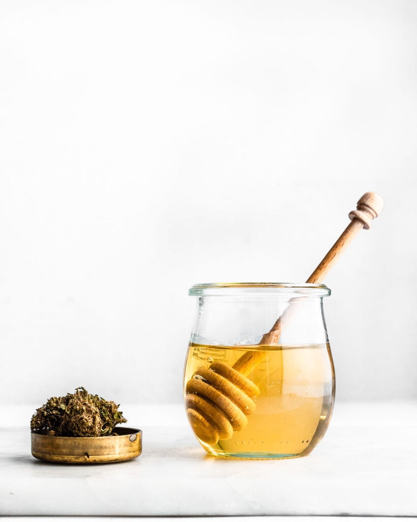 Cannabis-infused honey made with a high-CBD hemp strain in a small jar next to high-CBD hemp flower.