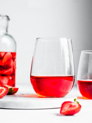 Two glasses of strawberry wine on a marble tray next to a large pitcher of strawberries soaking in white wine