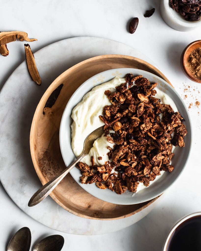 chocolate granola in a bowl of yogurt on a wooden plate, garnished with cacao and reishi mushroom