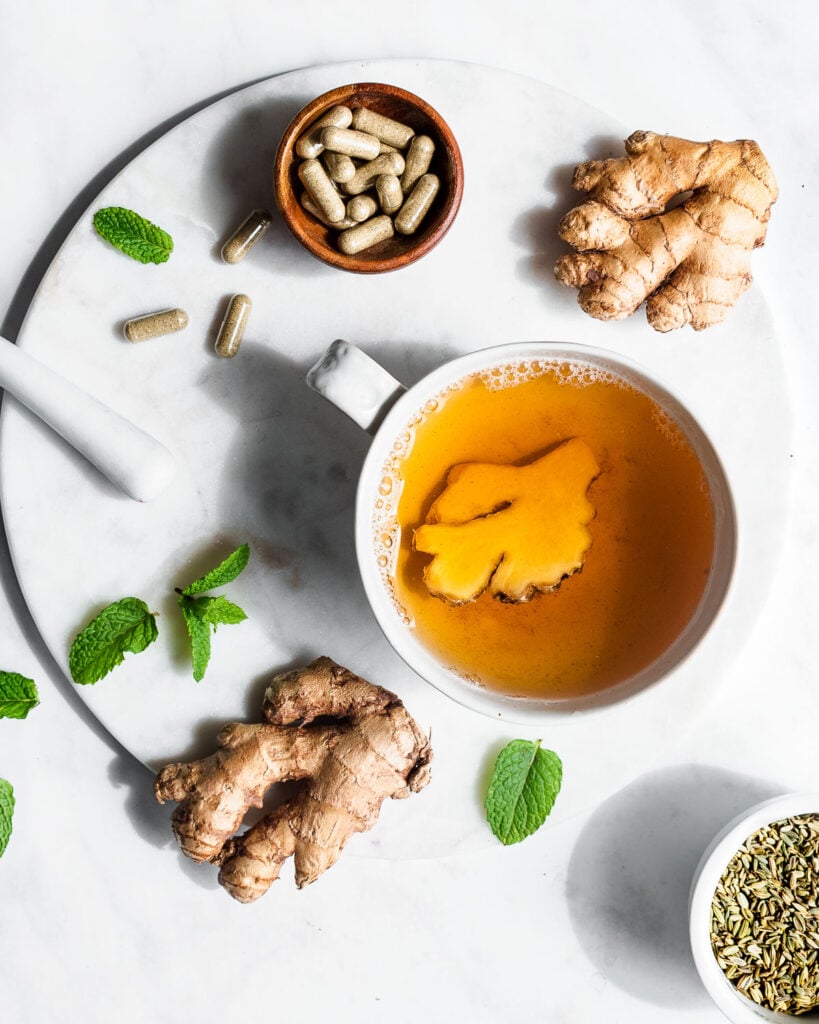 Ginger, mint leaves, fennel seed and herbal supplements on a marble surface next to a cup of ginger tea.