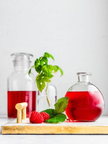 Raspberry liqueur in a round bottle on top of a marble serving tray next to raspberry leaves and fresh raspberries