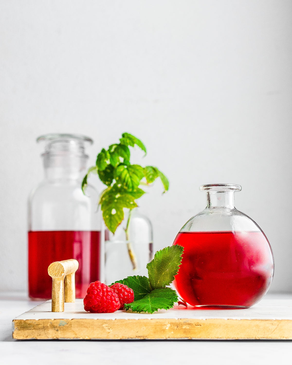 Raspberry liqueur in a round bottle on top of a marble serving tray next to raspberry leaves and fresh raspberries