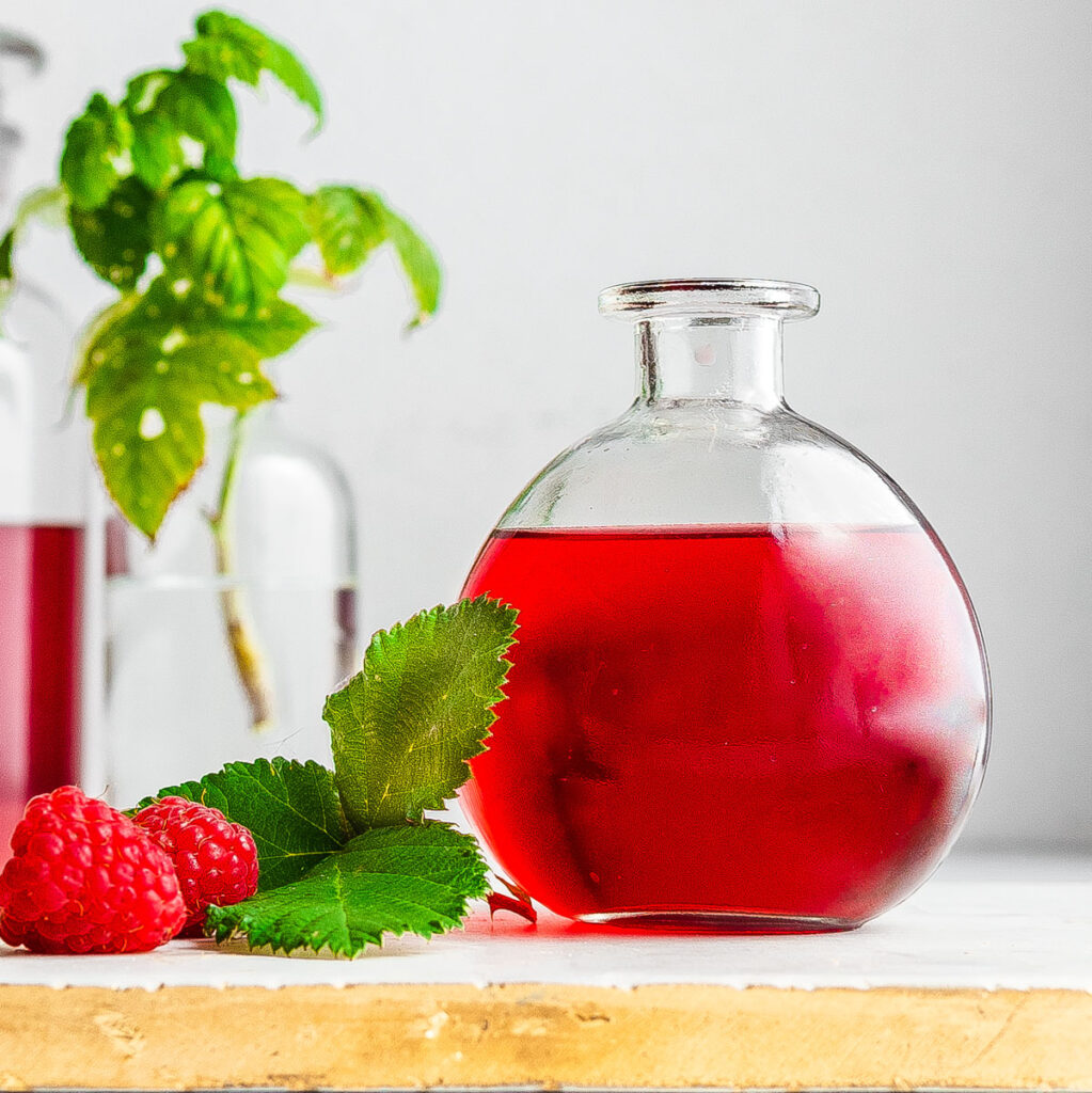 Raspberry liqueur in a round bottle on top of a marble serving tray next to raspberry leaves and fresh raspberries