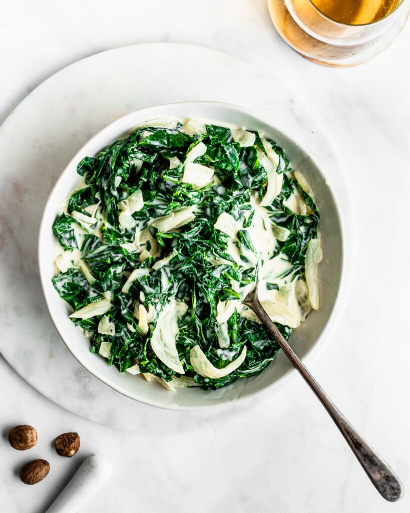 Creamed collard greens in a white bowl with a silver serving spoon on a marble background