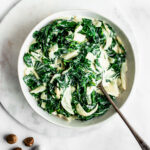 Creamed collard greens in a white bowl with a silver serving spoon on a marble background