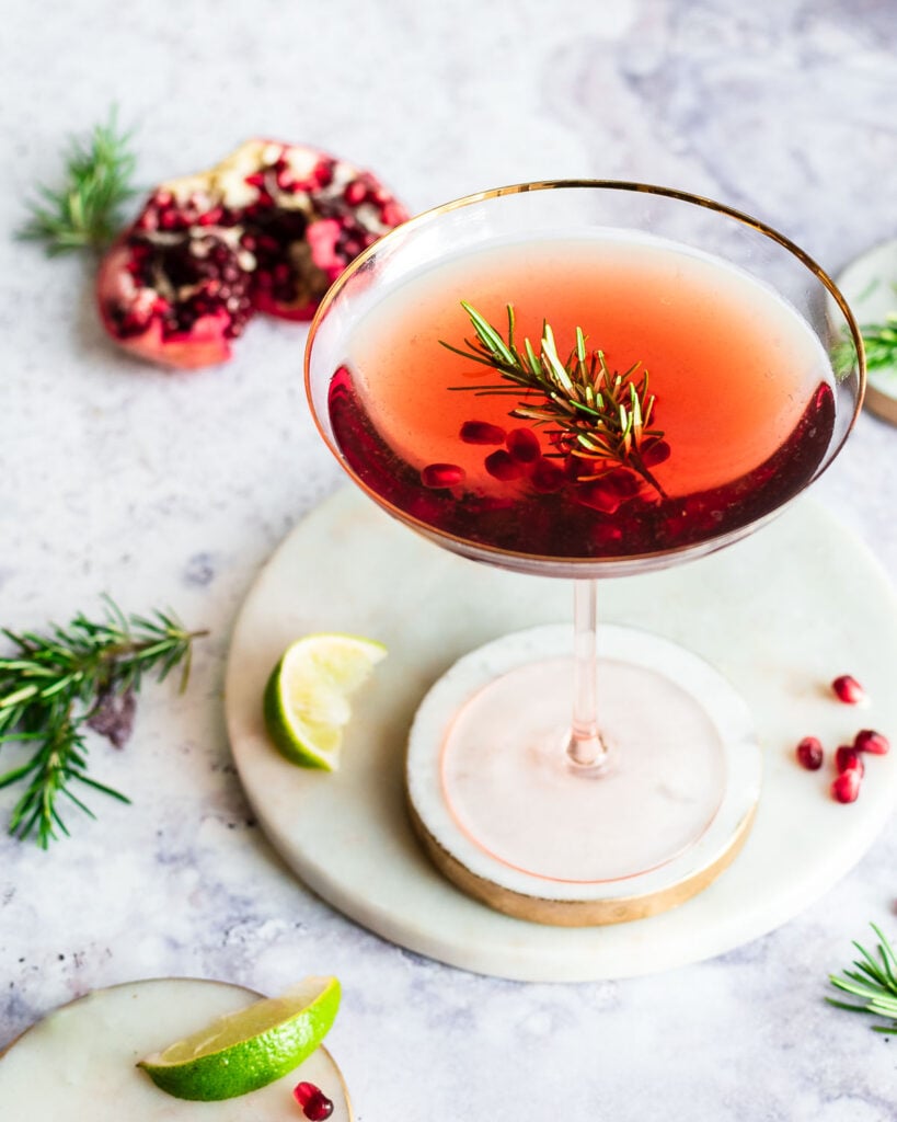 Pomegranate spritzer in a coupe glass, garnished with fresh rosemary and pomegranate arils; fresh pomegranate, lime wedge and rosemary branches in background.