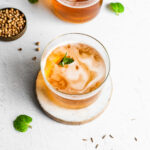 A glass of bread kvass garnished with fresh mint, fresh mint, coriander seeds and caraway seeds in background