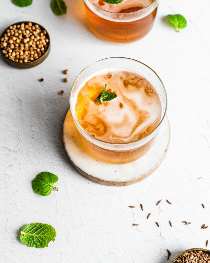 A glass of bread kvass garnished with fresh mint, fresh mint, coriander seeds and caraway seeds in background