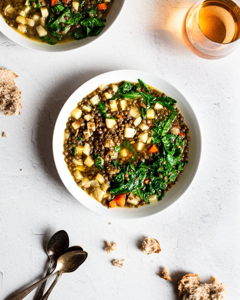 Two bowls of lentil stew on a white surface. Bread crumbs are on the table as well as a glass of beer.