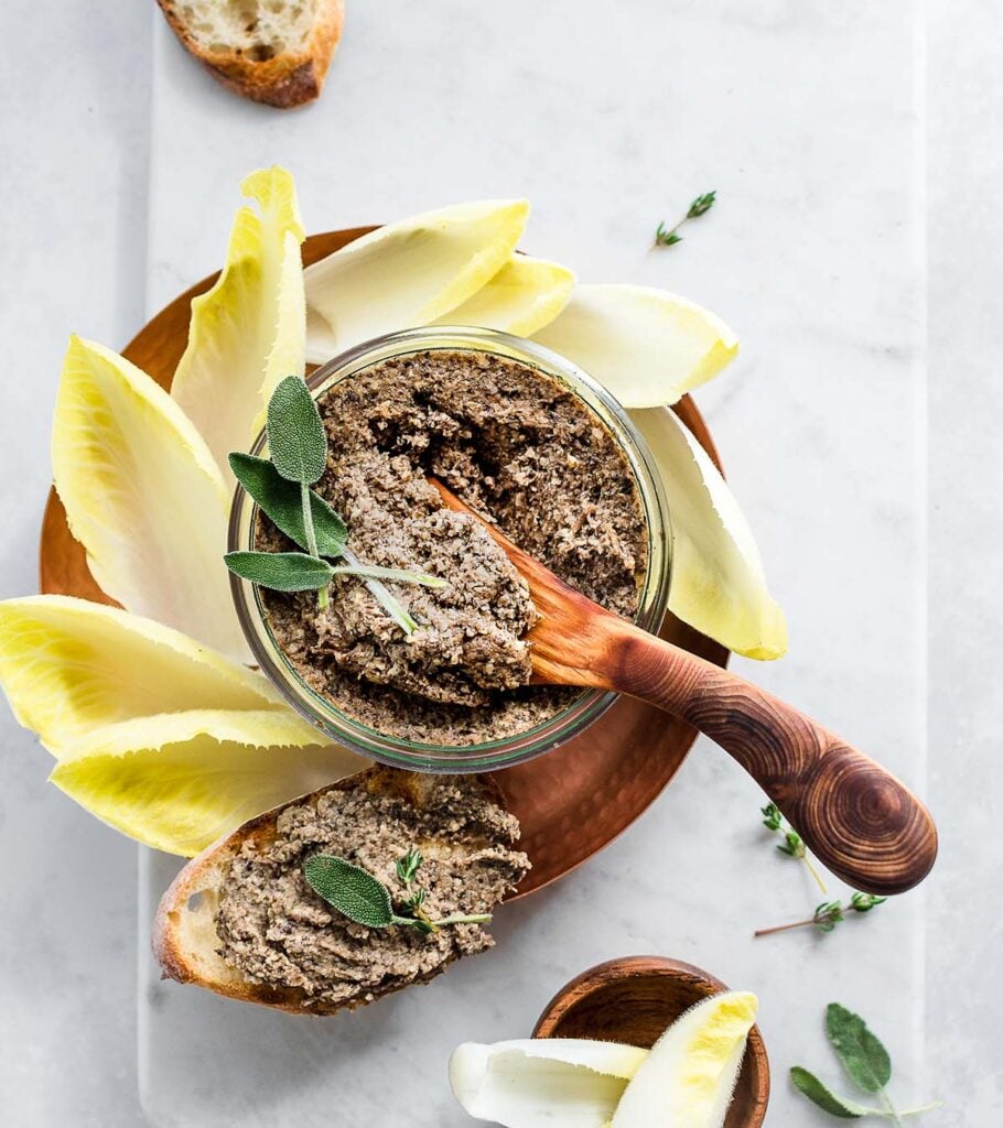 Mushroom pate in a glass jar on a copper dish garnished with sage leaves and fresh thyme, served with toasted bread and Belgian endive