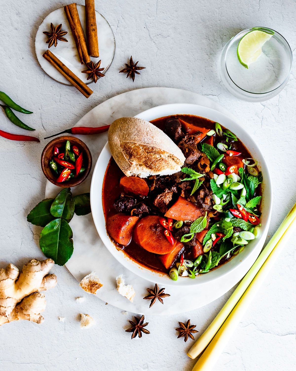 Bowlful of Bo Kho (Vietnamese Beef Stew) garnished with green onions, mint Thai basil, Thai chilies, and a baguette. Herbs and spices in background.