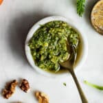 Carrot top pesto in a small bowl on a white surface, surrounded by walnuts and carrots