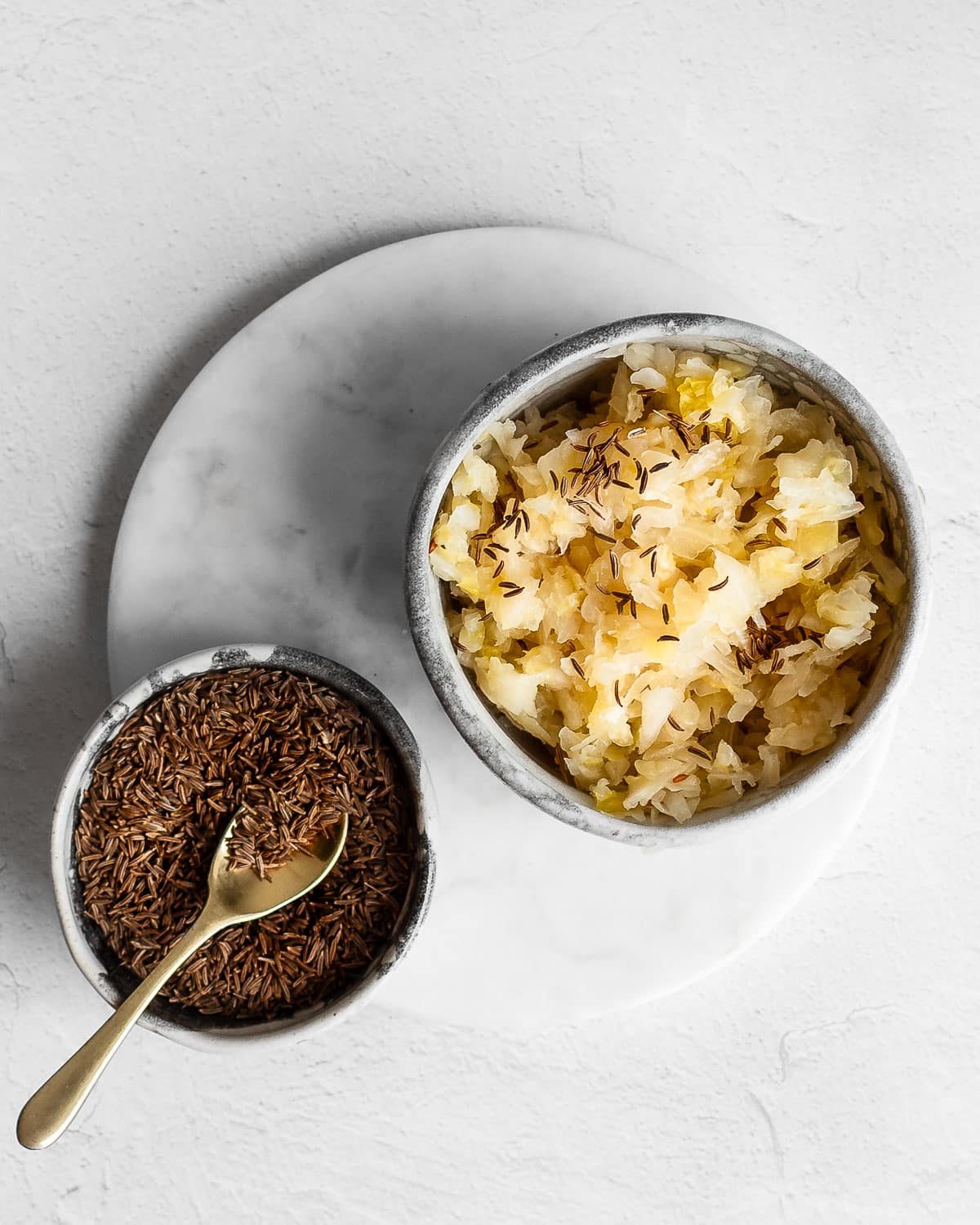 Caraway sauerkraut in a stoneware bowl on a marble surface next to a bowl of caraway seeds