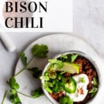Bison chili in a stoneware bowl garnished with jalapenos, sour cream, green onions, and lime