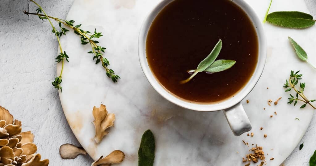 Herbal sipping broth in a white mug, herbs and mushrooms in the background