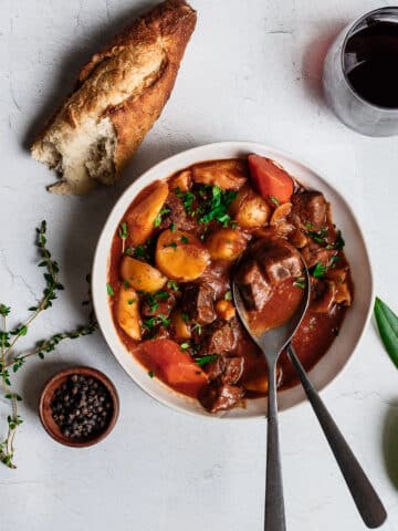 Bison stew in a white bowl, garnished with bay leaf, thyme and a baguette