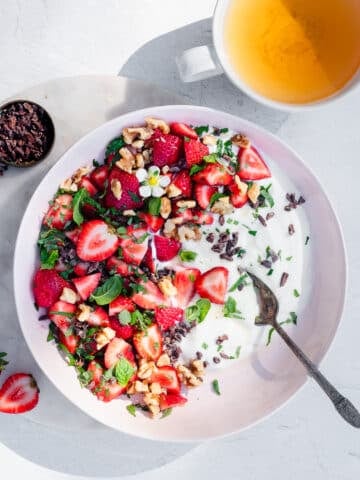 A pink bowl filled with yogurt, strawberries, mint, lemon balm, walnuts and cacao nibs